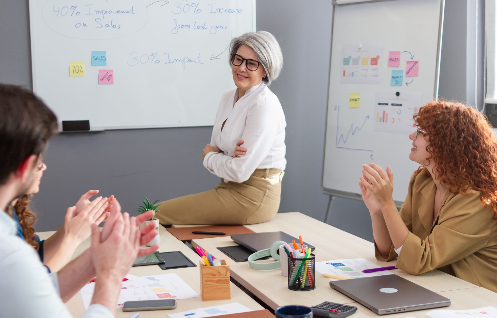 Coworkers clapping for mature businesswoman sitting on desk during a meeting